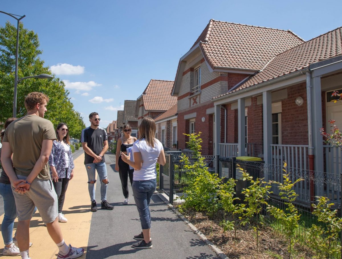 Une femme guide devant une maison en brique rouge qui explique à des visiteurs l'évolution de l'habitat minier des cités du Nors-Pas-de-Calais
