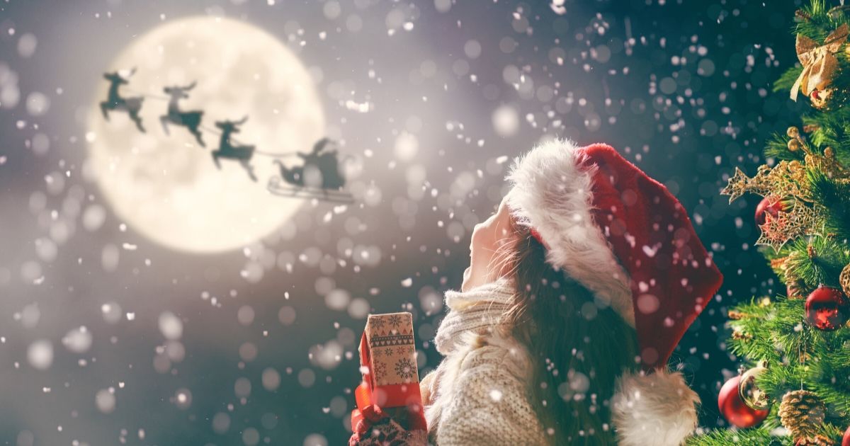 Une petite fille avec un bonnet de noël et un cadeau à la main sous la neige et qui regarde le traineau du père noël dans le ciel